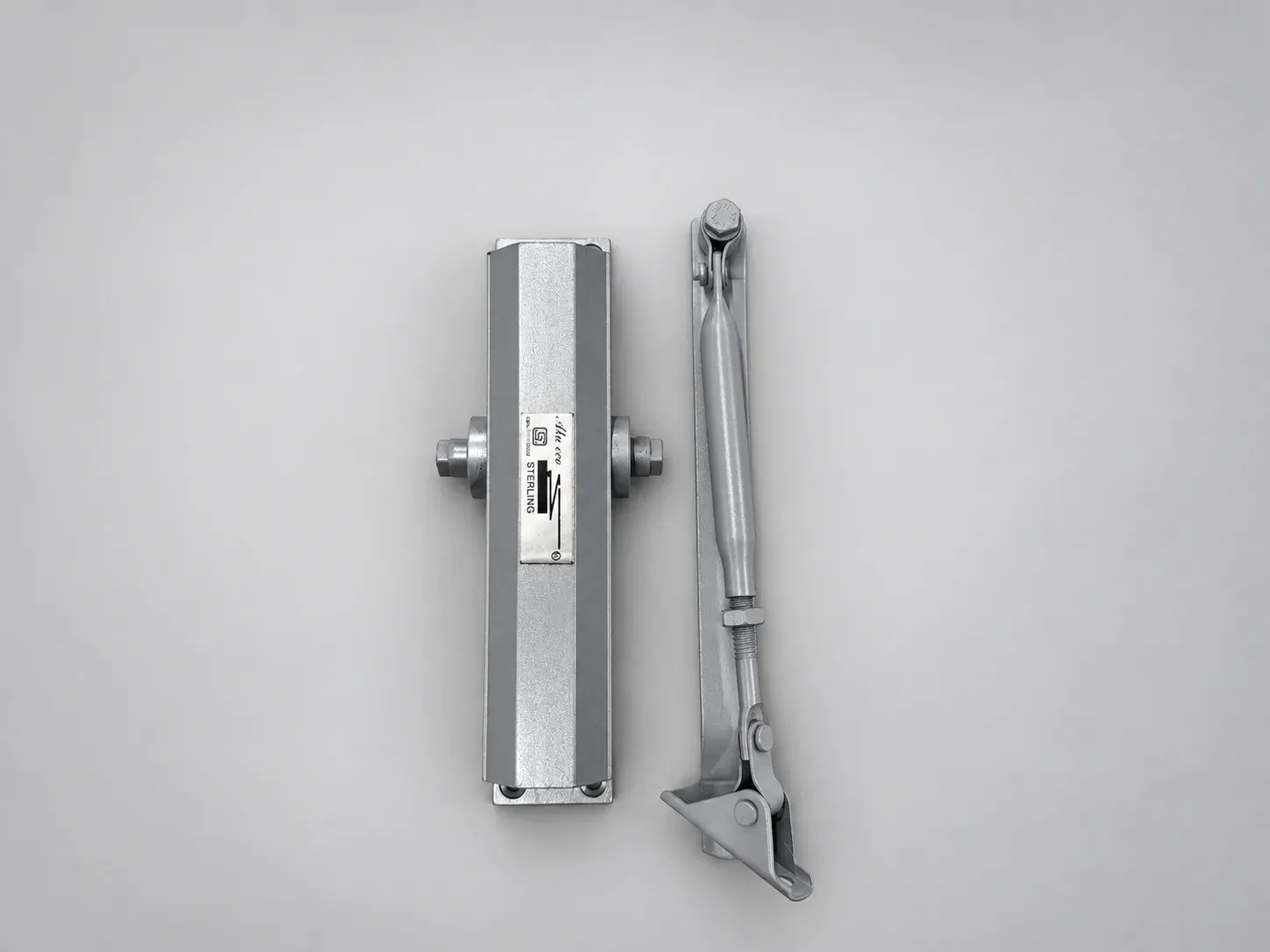 Sterling DC 1500 Alu Eco door closer body with its Alu Eco label plate, laid beside its lever arm, silver, overhead view, studio photograph
